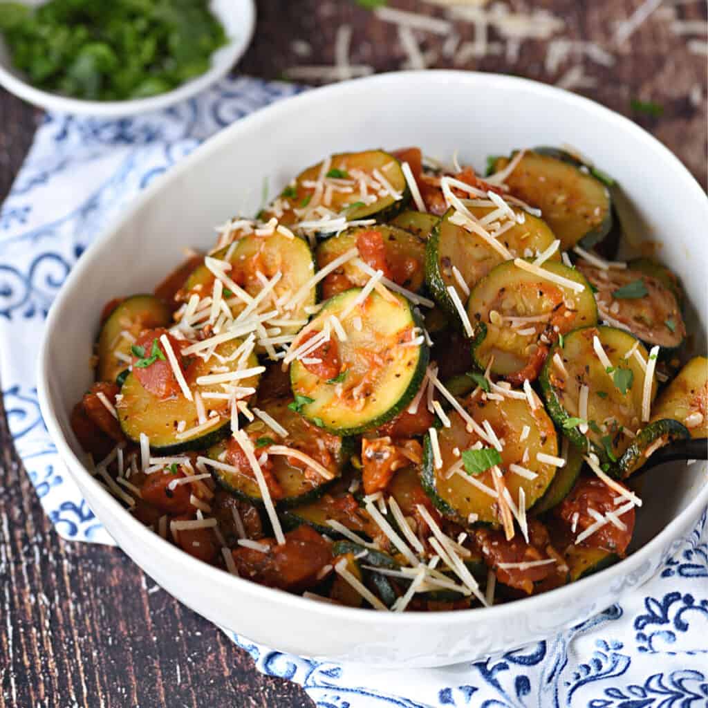 a large white bowl filled with Sauteed zucchini slices and diced tomatoes topped with grated Parmesan cheese and chopped parsley.