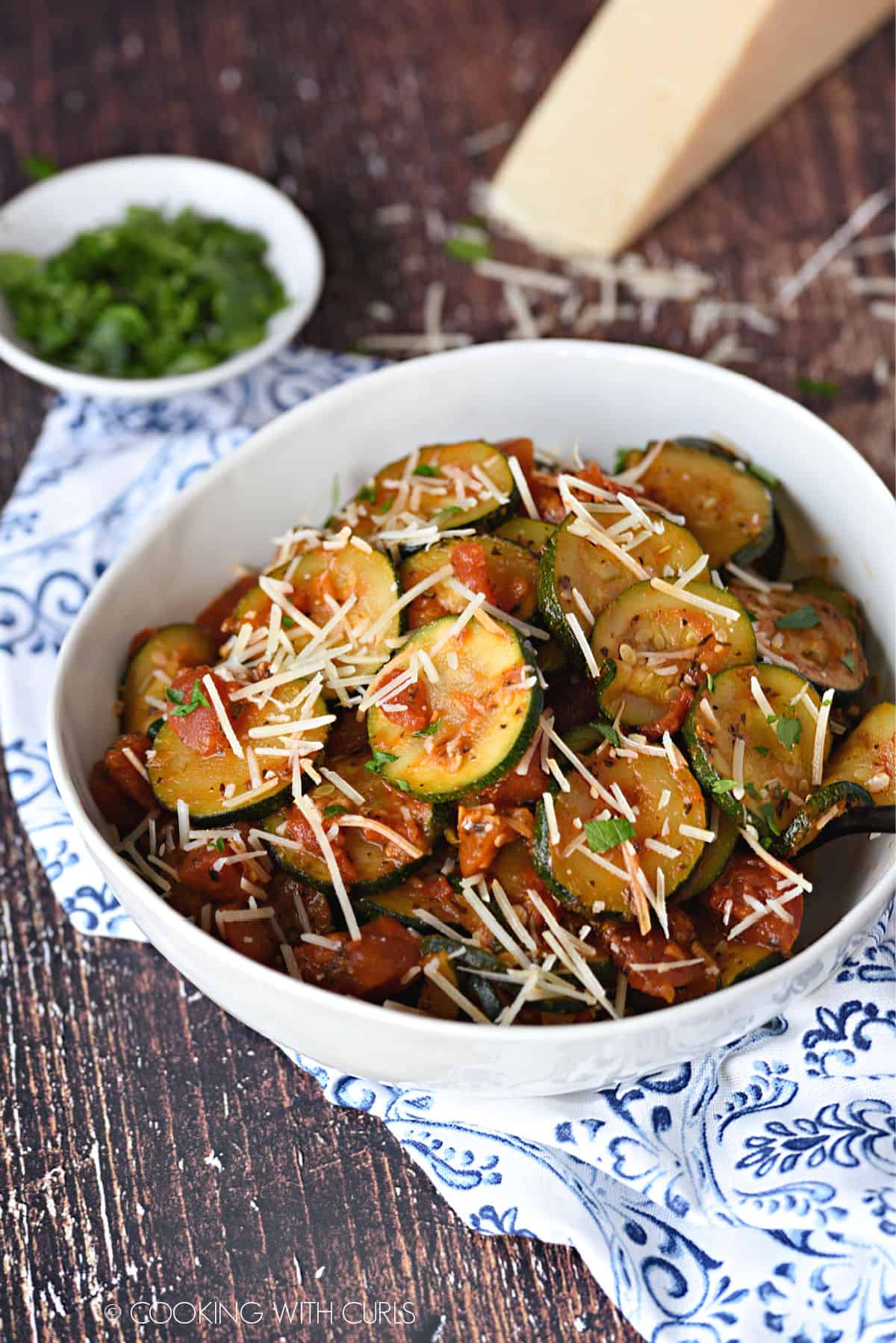a large white bowl filled with Sauteed zucchini slices and diced tomatoes topped with grated Parmesan cheese and chopped parsley.