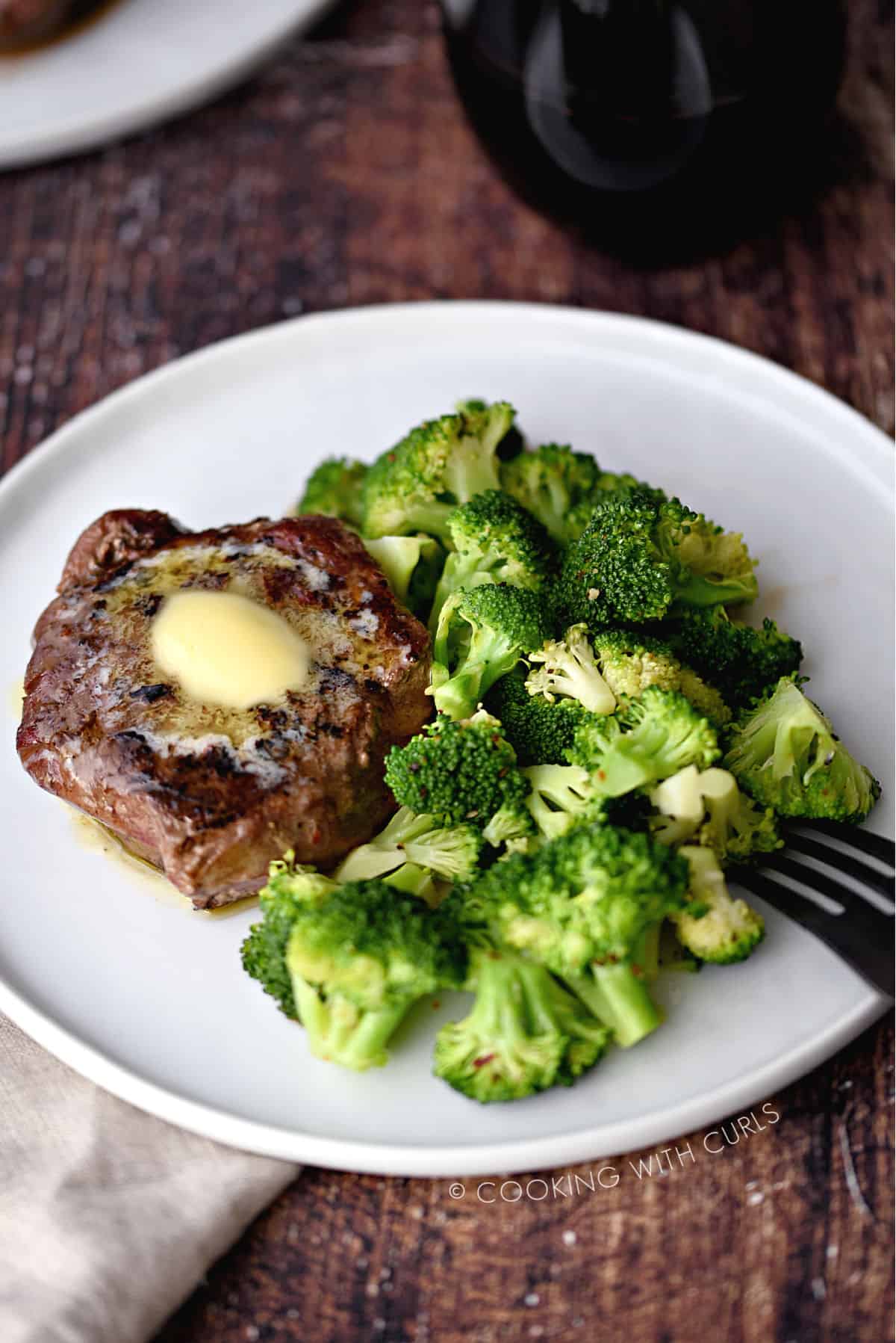 steamed broccoli and a butter topped steak on a white plate with a black fork on the edge.