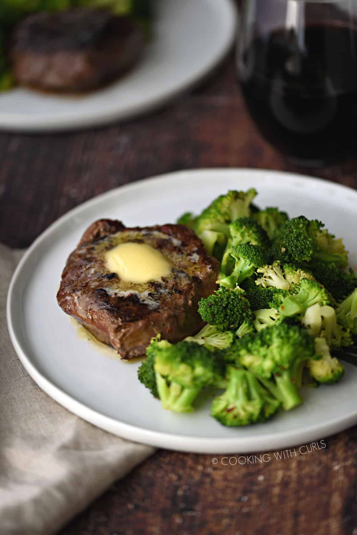 a butter topped filet next to a pile of broccoli florets on a white plate, with a glass of red wine and a second plate in the background.