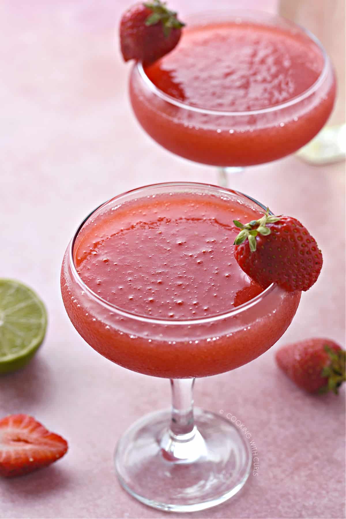 Two coupe glasses filled with frozen strawberry daiquiri and garnished with a small strawberry on the rim. A cut and whole strawberry are sitting at the base with a lime half peaking out through the edge.