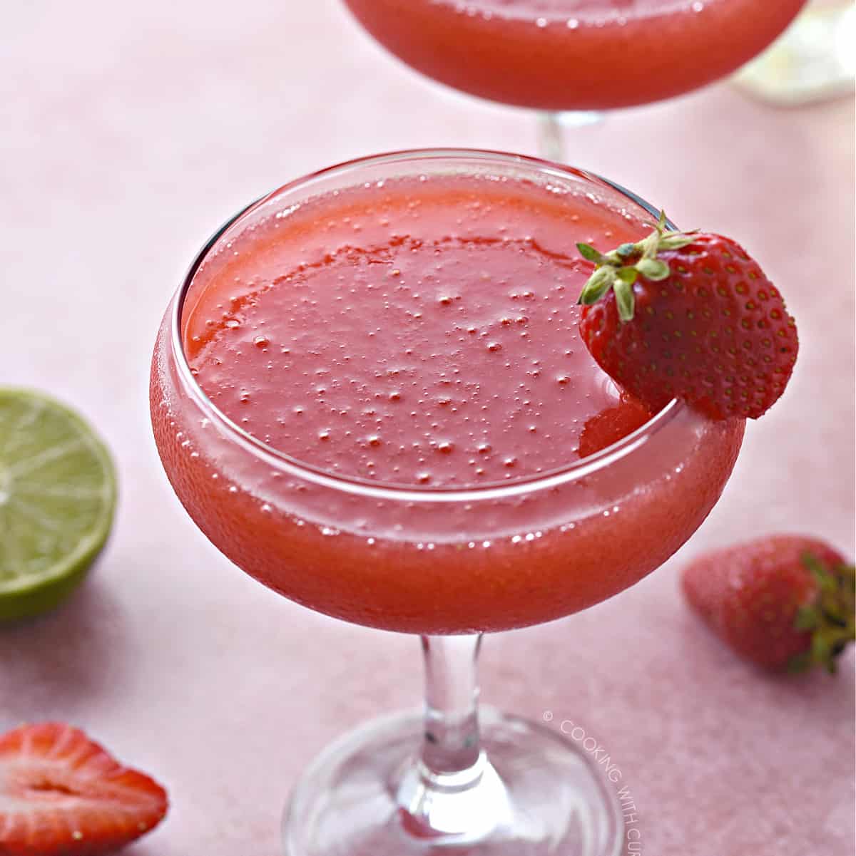 a close up image of a frozen strawberry daiquiri in a coupe glass with a whole strawberry stuck onto the rim of the glass.