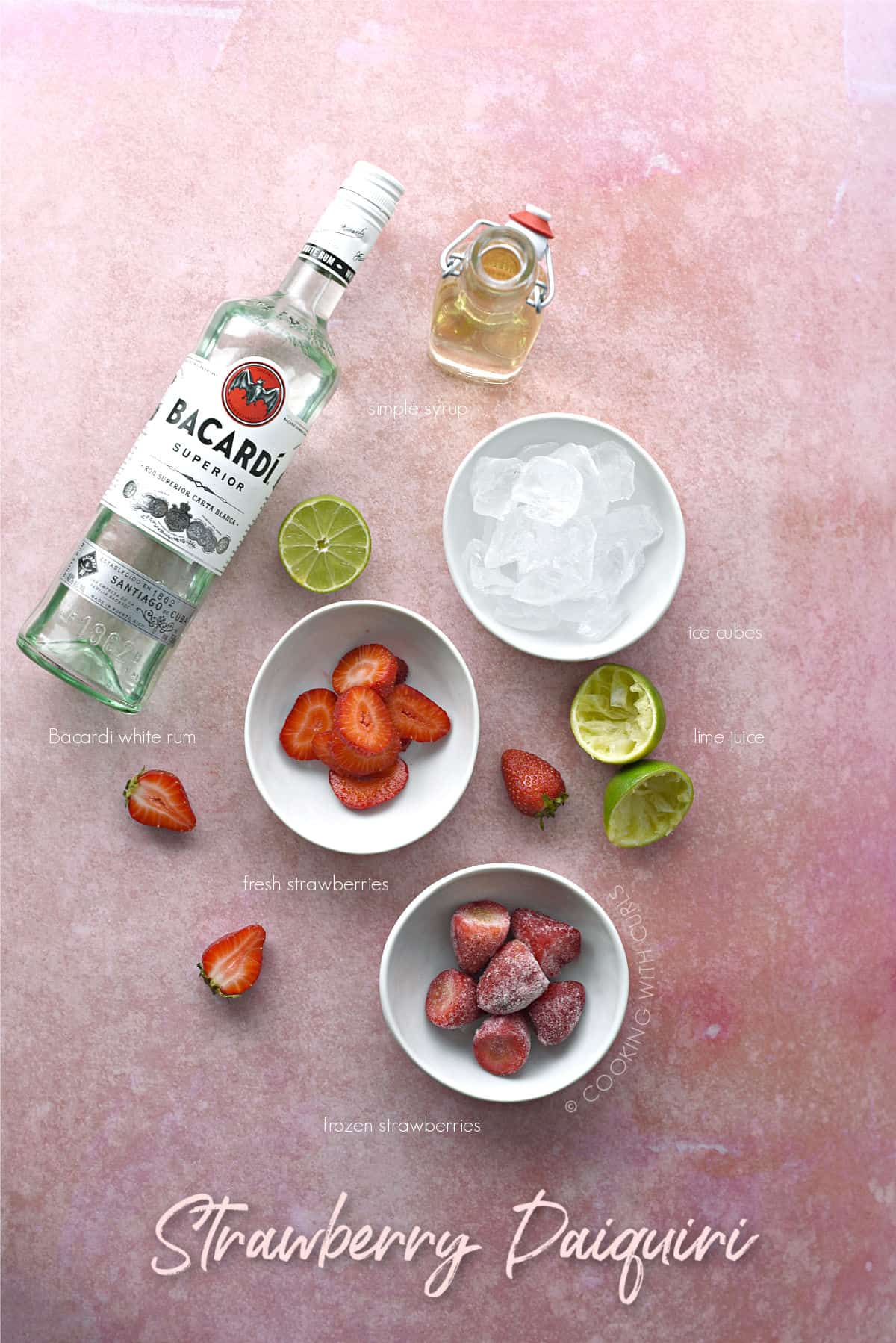 a bottle of Bacardi rum laying on it's side next to a bottle of simple syrup, and bowls of strawberries and ice cubes with squeezed limes in between. 