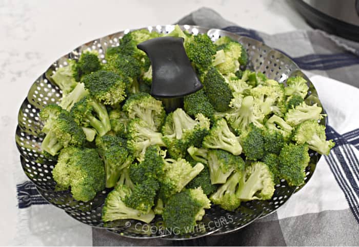 fresh broccoli florets in a metal steamer basket sitting on a blue and white checkered towel. 