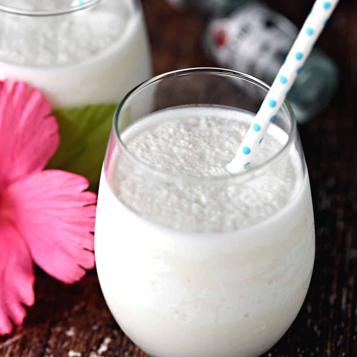 A creamy Keto Pina Colada with Coconut Milk in a glass with a blue polka dot straw with a second cocktail and a pink flower in the background.