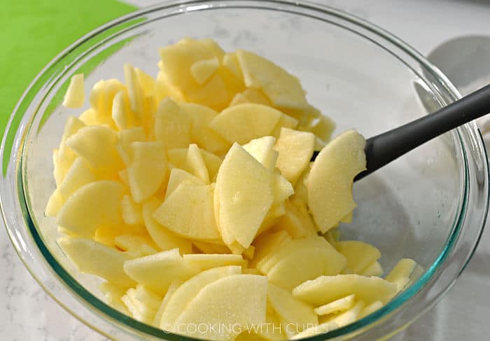 Apple slices tossed with sugar in a clear glass bowl with a gray rubber spoon. 