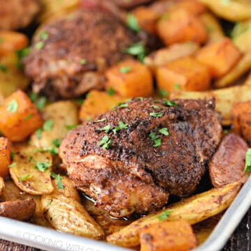 a close-up image of two chicken thighs surrounded by potatoes and squash cubes on a sheet pan.