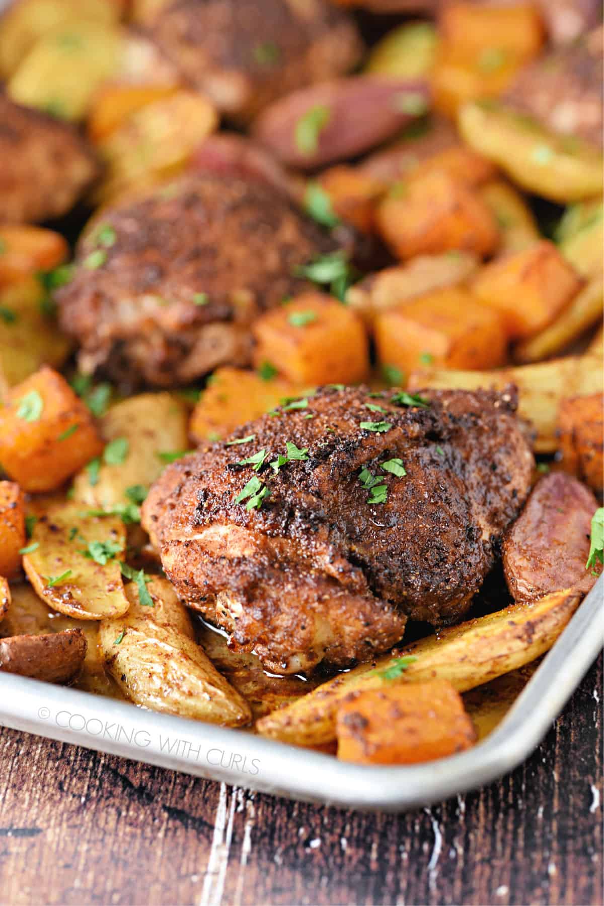 a close-up image of spice rubbed chicken thighs surrounded by roasted potatoes and butternut squash on a metal sheet pan.