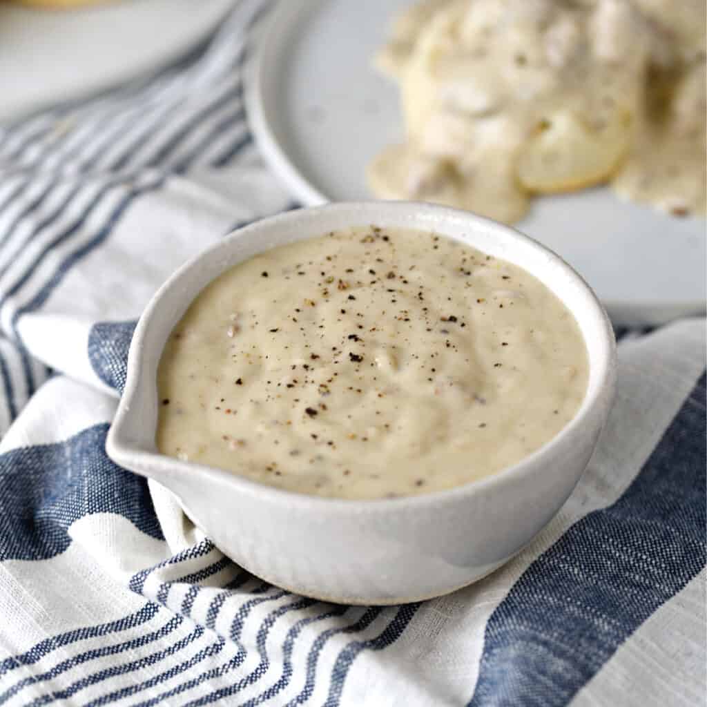 thick, creamy sausage gravy in a white serving bowl with a plate of biscuits and gravy in the background.