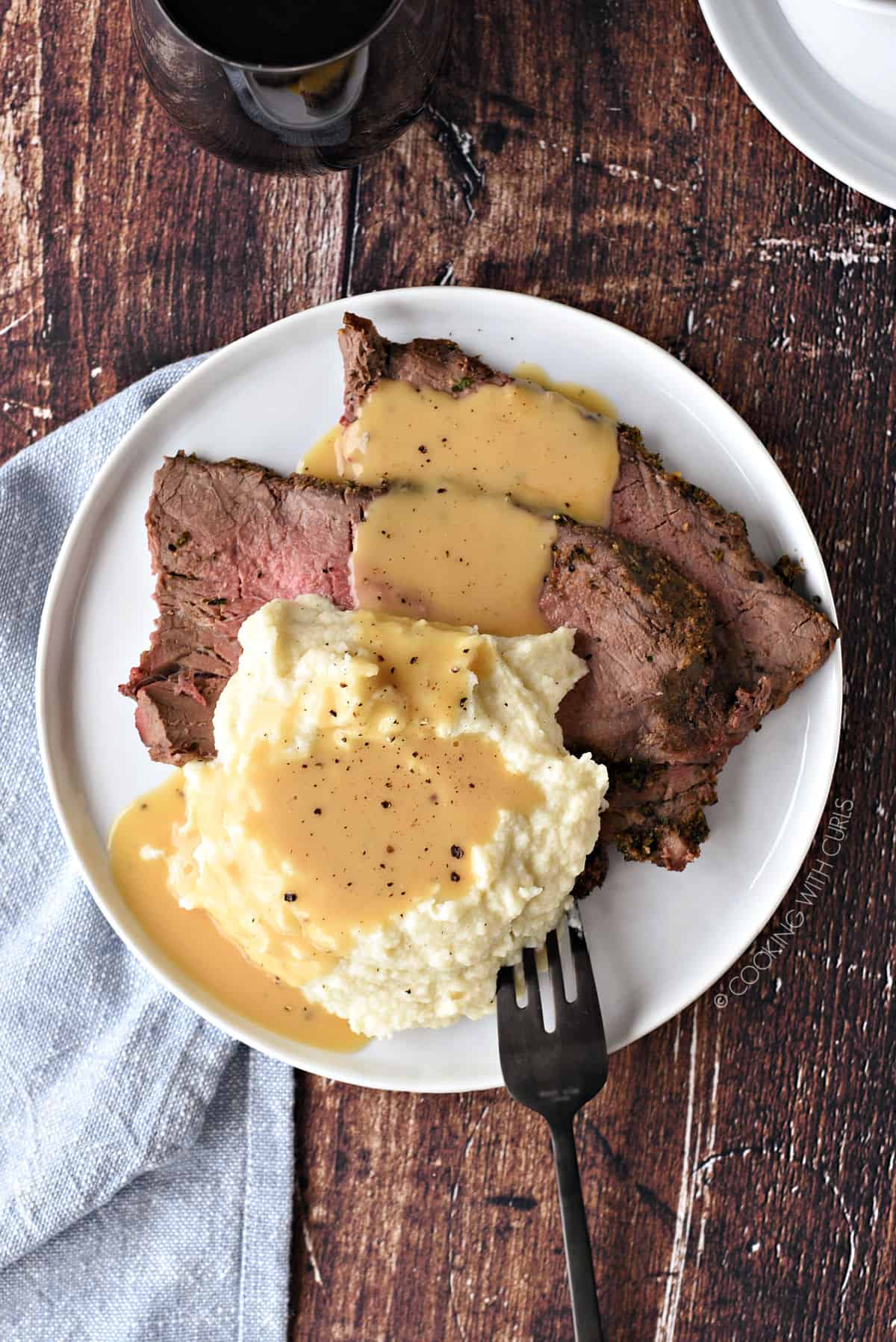 looking down on a plate of sliced roast beef and mashed cauliflower topped with beef gravy and ground black pepper, with a blue napkin under the plate.