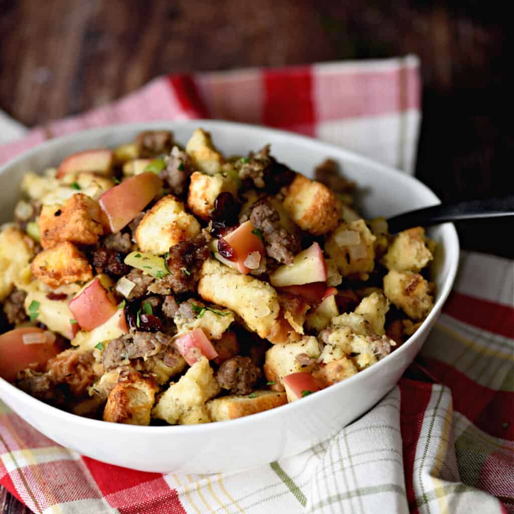 a closeup image of a white bowl filled with sausage, apple and cranberry stuffing sitting on a red plaid napkin.