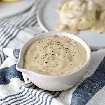 thick, creamy sausage gravy in a white serving bowl with a plate of biscuits and gravy in the background.