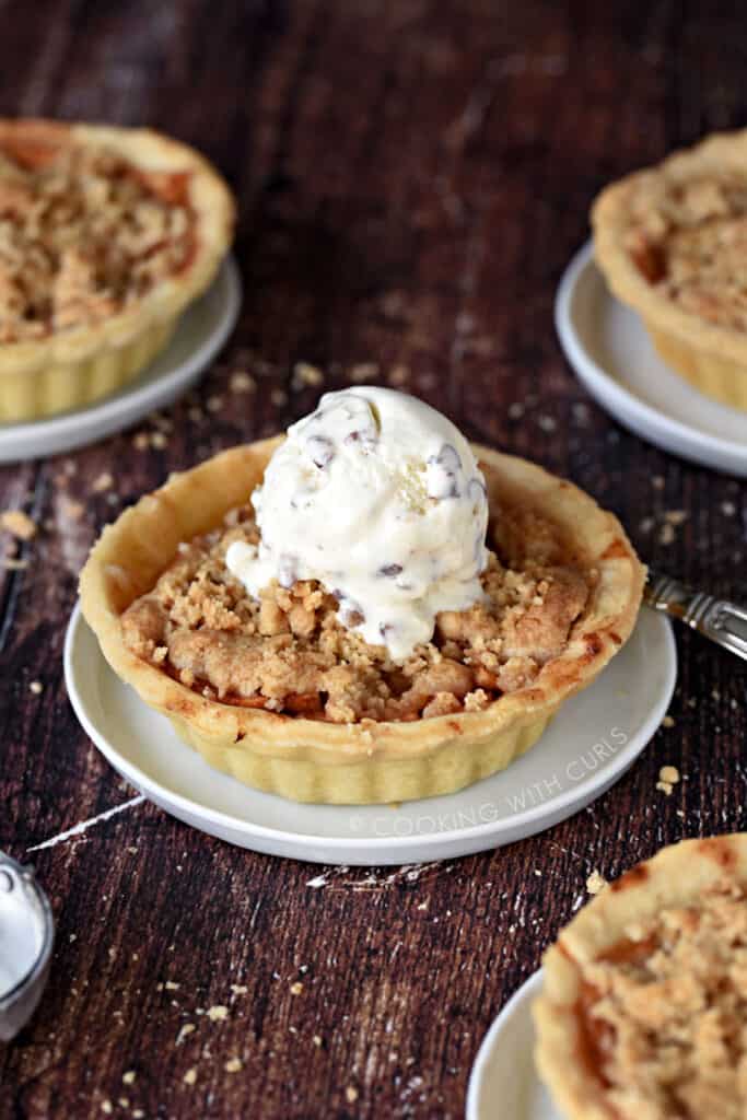 An ice cream topped Mini Dutch Apple Pie in the center of a wooden surface, surrounded by three additional mini pies on small white plates.