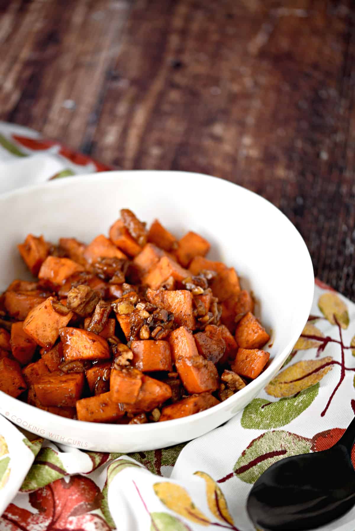 yam cubes tossed with pecans, butter, sugar and spices in a white bowl that is sitting on a fall leaves napkin.