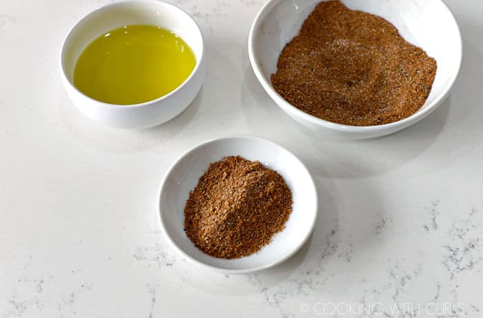 a small white bowl with olive oil, a bowl with spices, and a smaller bowl with a tablespoon of the spice mixture. 