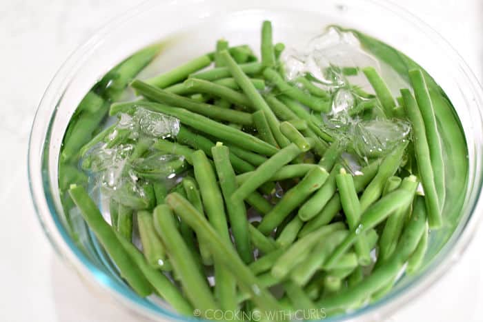 blanched green beans in a glass bowl filled with ice cubes and water. 