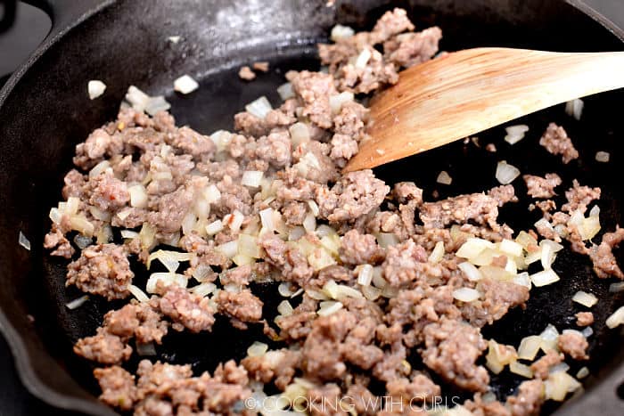 ground sausage and diced onions cooking in a cast iron skillet with a bamboo turner in the upper right hand corner. 