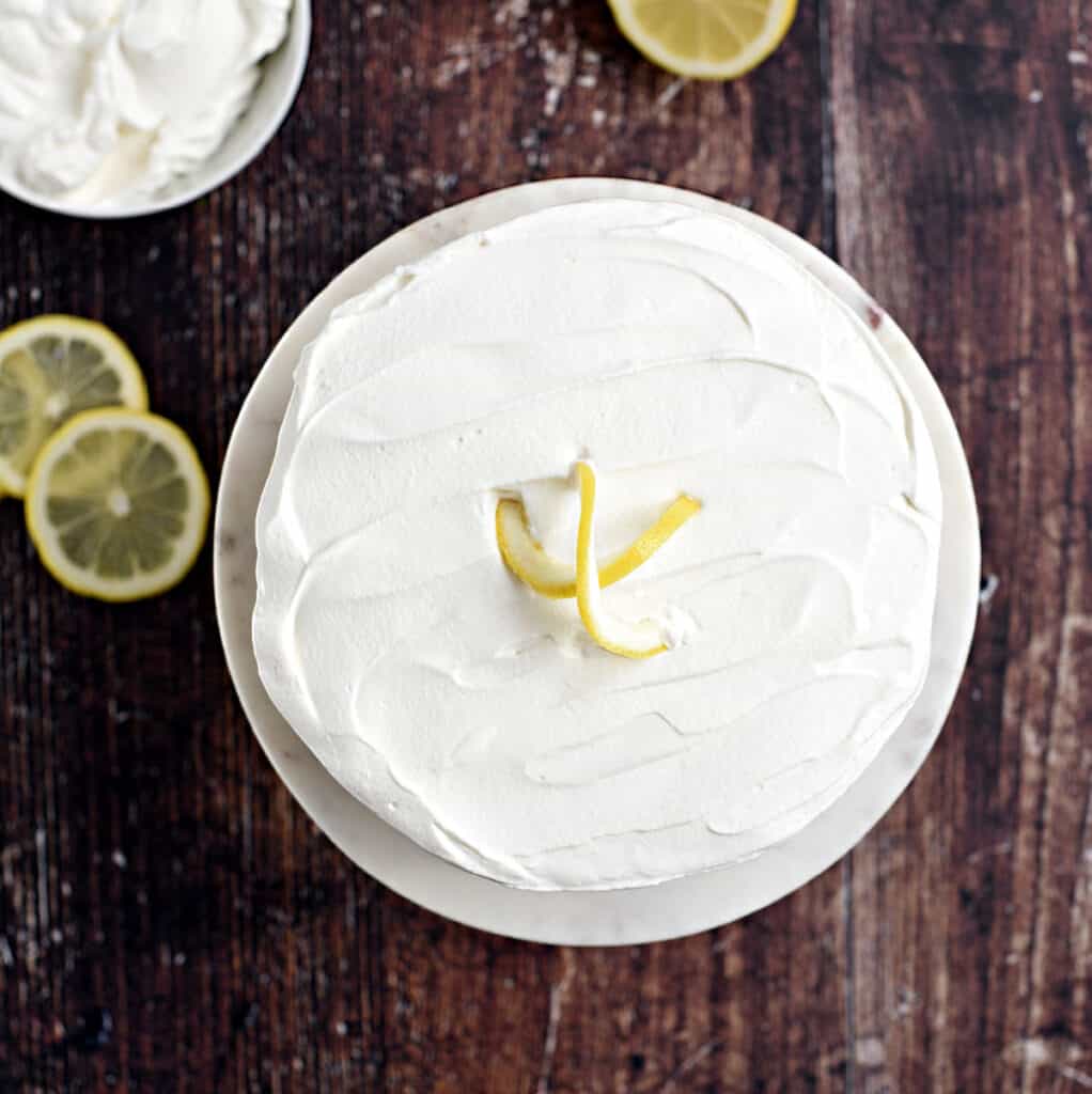 looking down on a whole lemon cheesecake topped with whipped cream, with a bowl of whipped cream and lemon slices in the background.