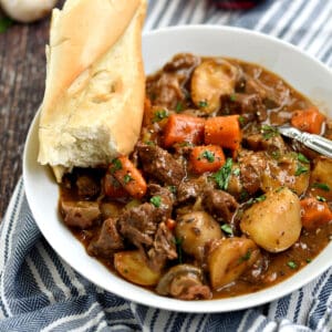 A big white bowl filled with Beef Bourguignon with a piece of baguette sitting on the side of the bowl.
