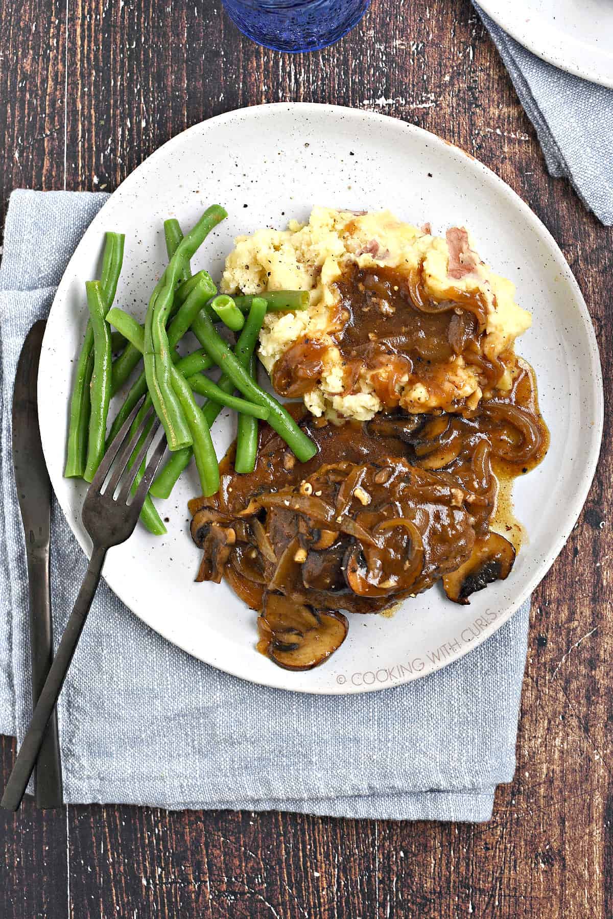 Looking down on a plate of mushroom gravy topped salisbury steak and mashed potatoes with green beans on the side.