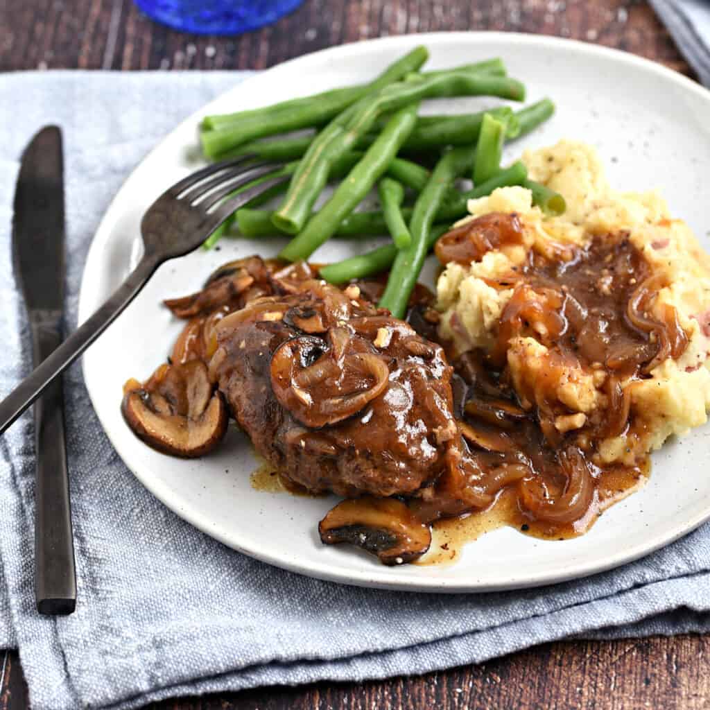 Salisbury Steak with mushroom gravy on a white plate with mashed potatoes and green beans, sitting on a blue napkin.