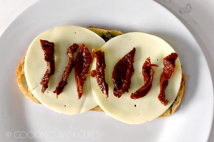 Looking down on strips of sun-dried tomatoes lined up on top of provolone cheese slices on a slice of bread sitting on a white plate. 