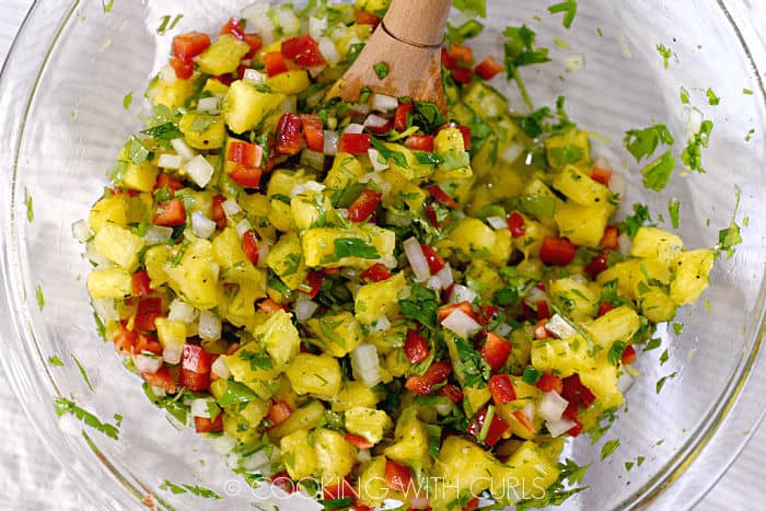 Pineapple salsa mixed together in a glass bowl with a wooden spoon. 