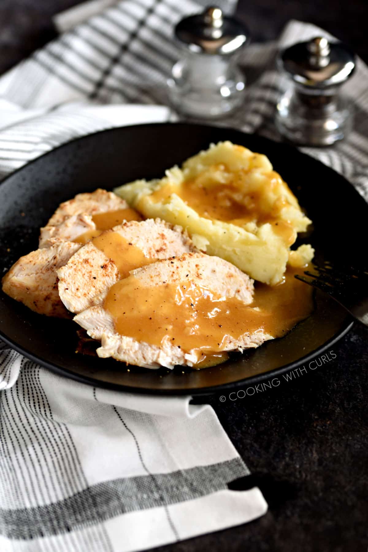 Turkey Gravy poured over sliced turkey and mashed potatoes on a black plate.