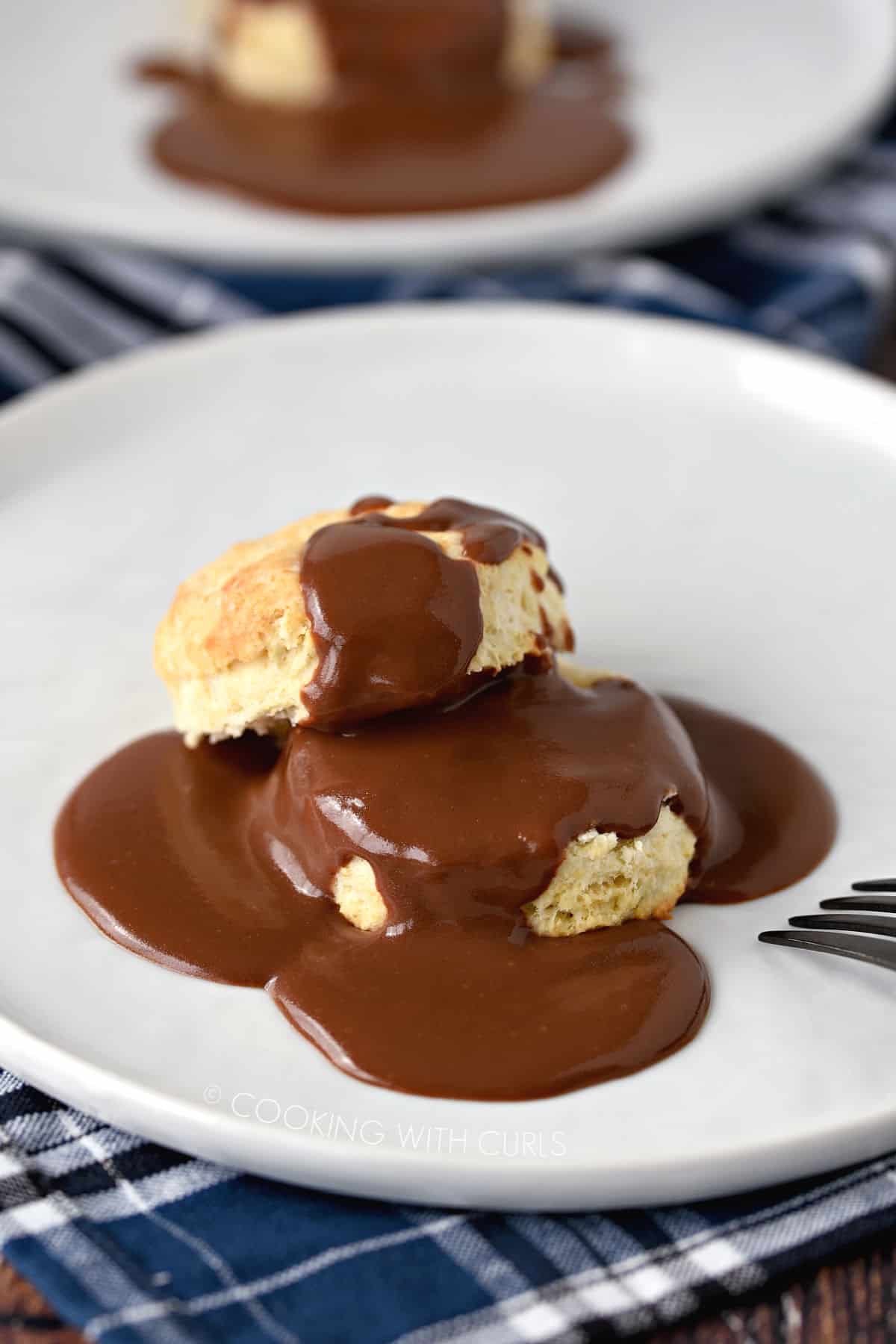 A biscuit split in half and covered with thick, chocolate gravy on a white plate with a second plate in the background.