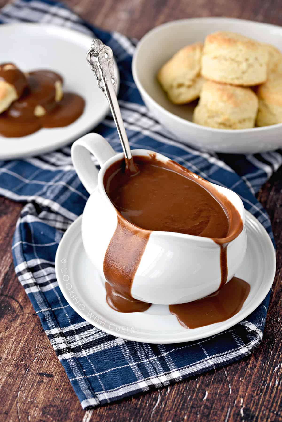 Chocolate Gravy overflowing a white gravy boat sitting on a white plate with a ladle sticking up out of the backside, with a place of chocolate gravy covered biscuits on the left and a bowl of biscuits in the upper right corner.
