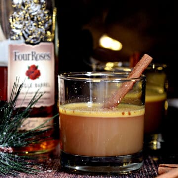 Two glasses of hot buttered bourbon with cinnamon sticks and a bottle of bourbon on the side.