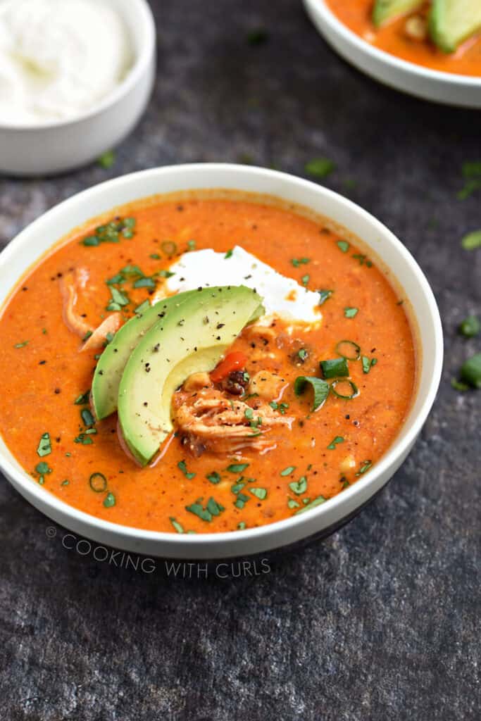 A bowl of creamy chicken enchilada soup topped with yogurt, avocado slices and diced green onions.