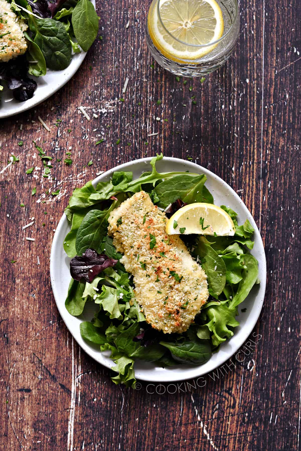 Looking down on a panko crusted fish filet on a bed of leafy greens with a lemon wedge on the side.
