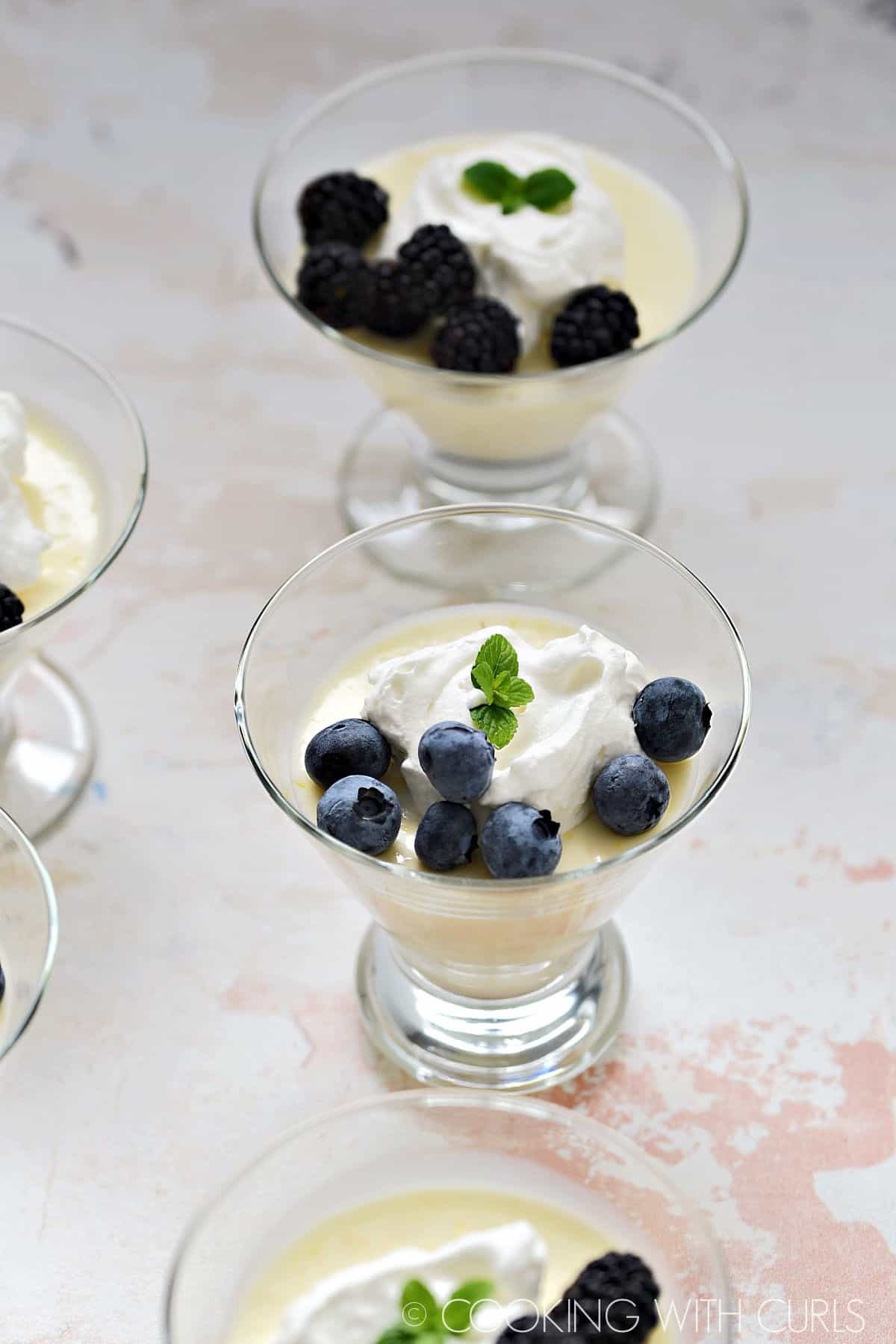 Four clear dessert glasses filled with lemon custard and topped with a dollop of whipped cream and a handful of blueberries.