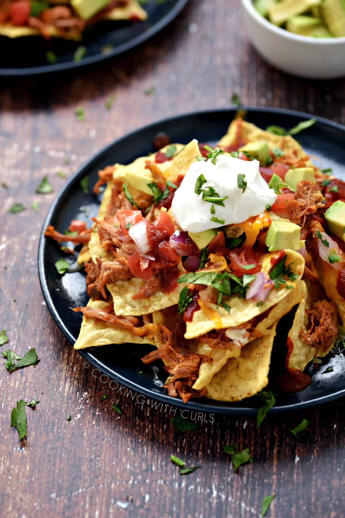 Tortilla chips, melted cheese, pulled pork, pico de gallo, avocado chunks, sour cream and cilantro piled up on a blue plate.