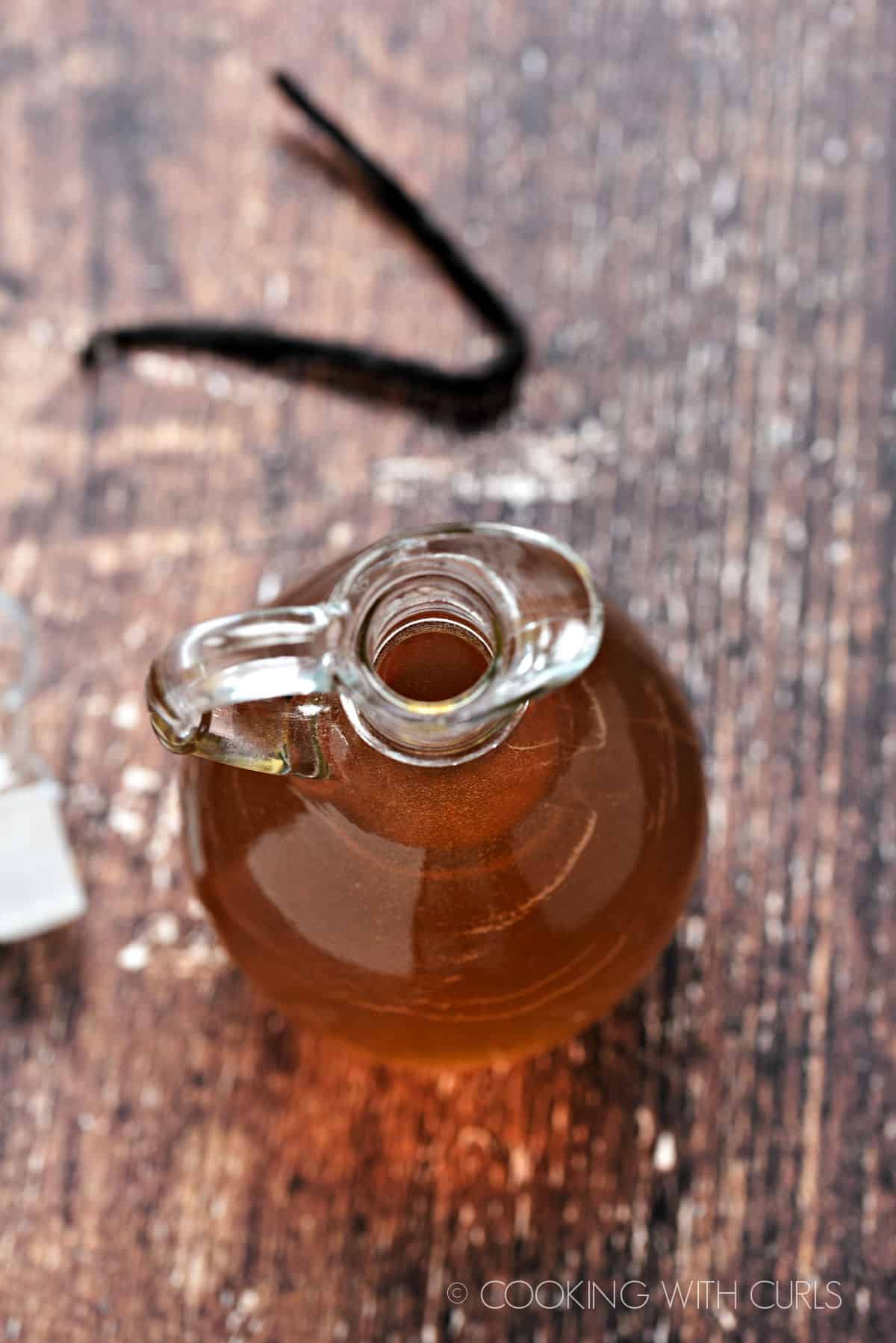 Looking down into a glass bottle with a spout filled with vanilla simple syrup with a vanilla bean laying in the upper left corner.