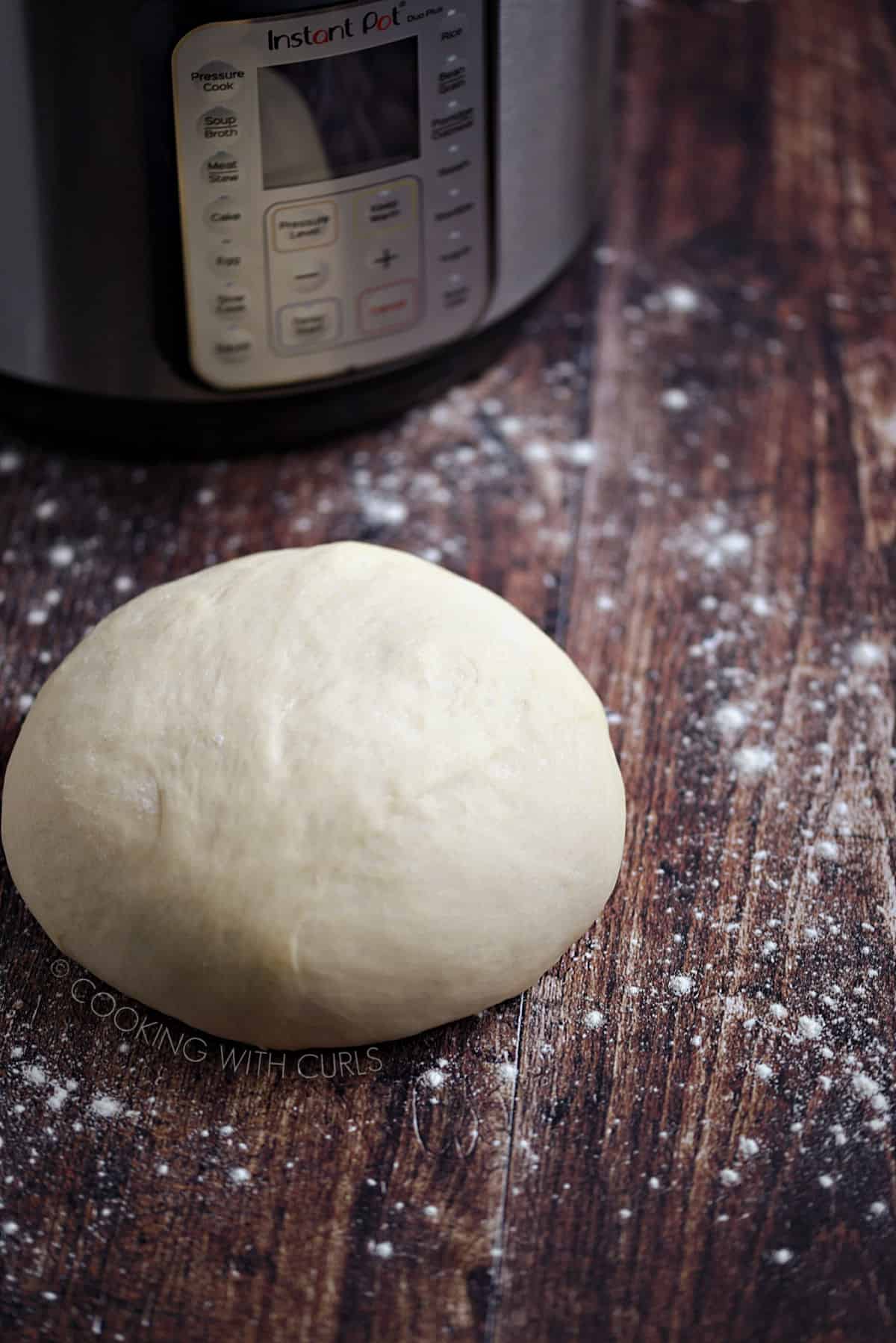 A ball of pizza dough on a flour dusted wood surface with an Instant Pot in the background.