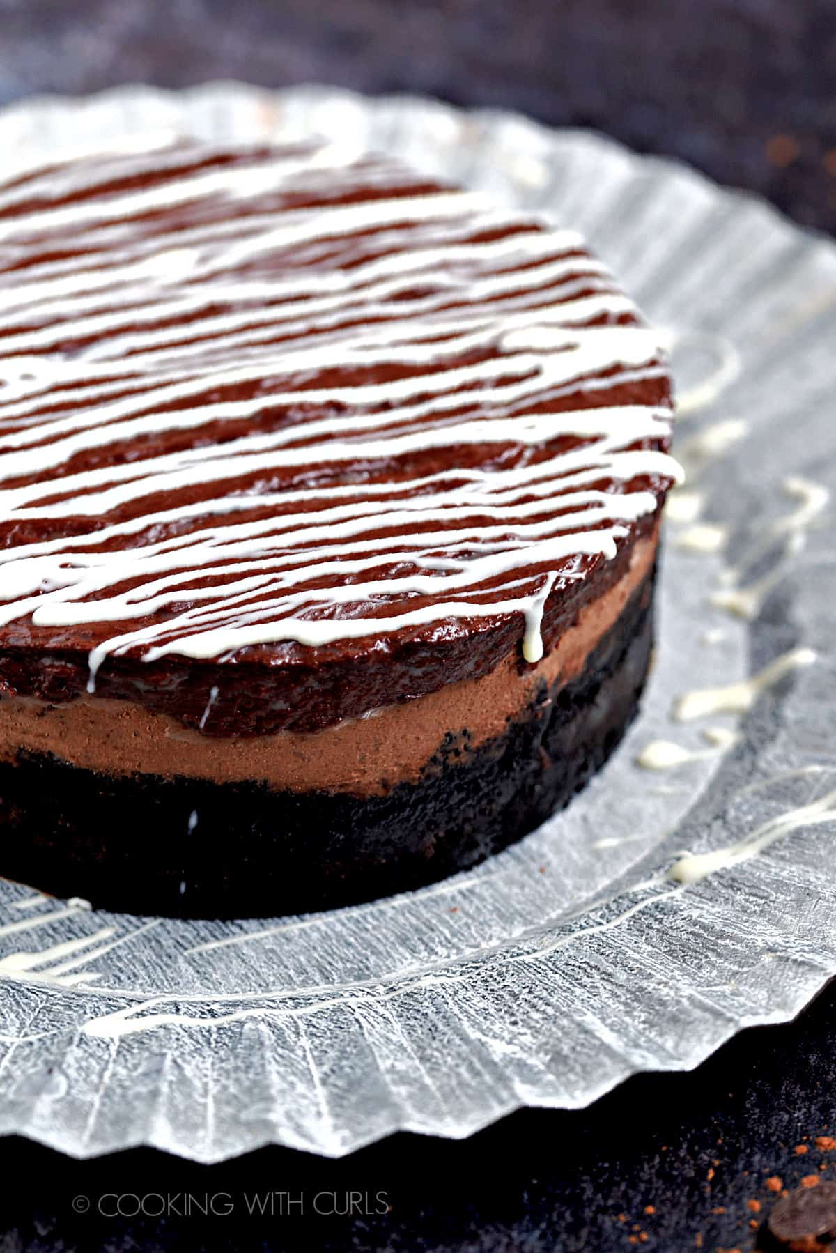 White chocolate drizzled over a chocolate cheesecake sitting on a ruffled metal serving platter.