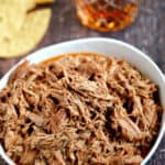pulled pork in a white bowl with tortilla chips and a glass of whiskey in the background.