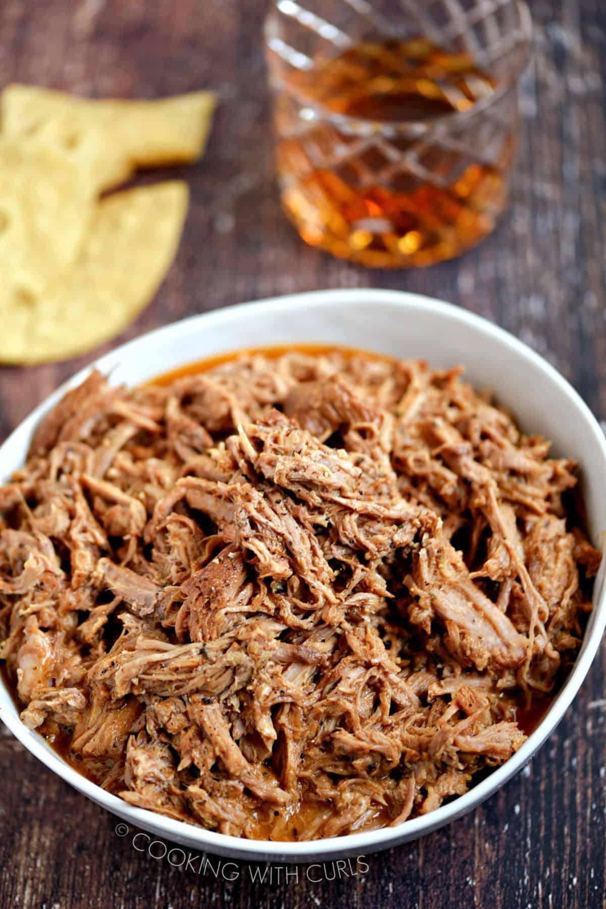 Pulled Pork in a white bowl with tortilla chips and a glass of whiskey in the background.