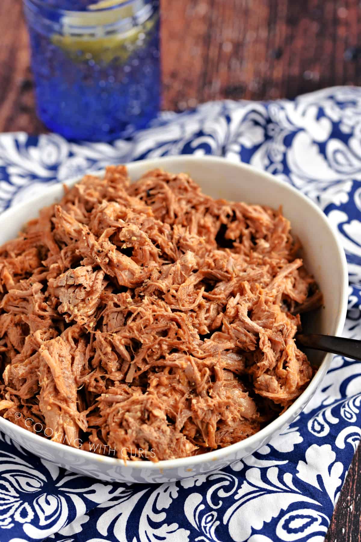 A large white bowl filled with Instant Pot Pulled Pork sitting on a blue and white designed napkin with a blue glass in the background.