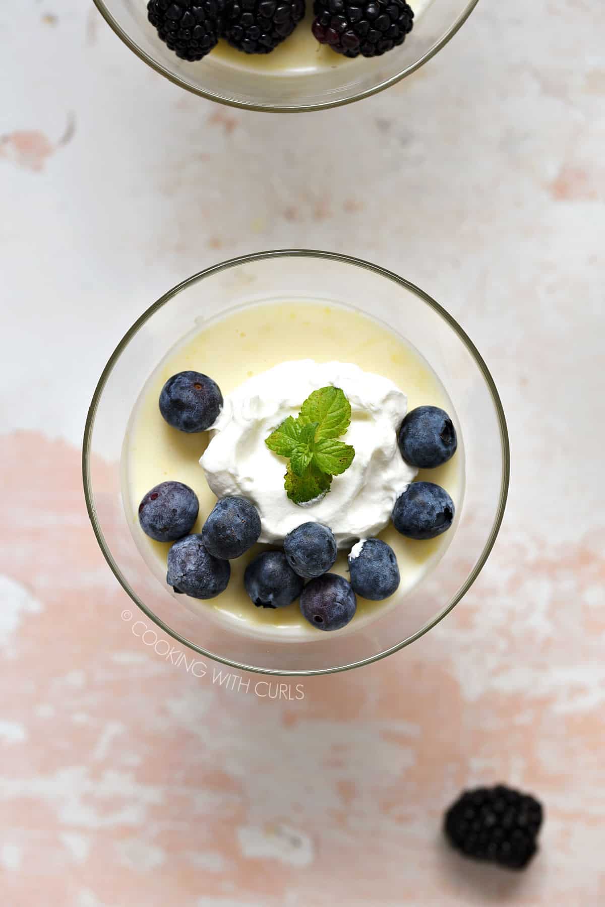 Looking down on a clear dessert glass filled with lemon custard and topped with whipped cream and blueberries.