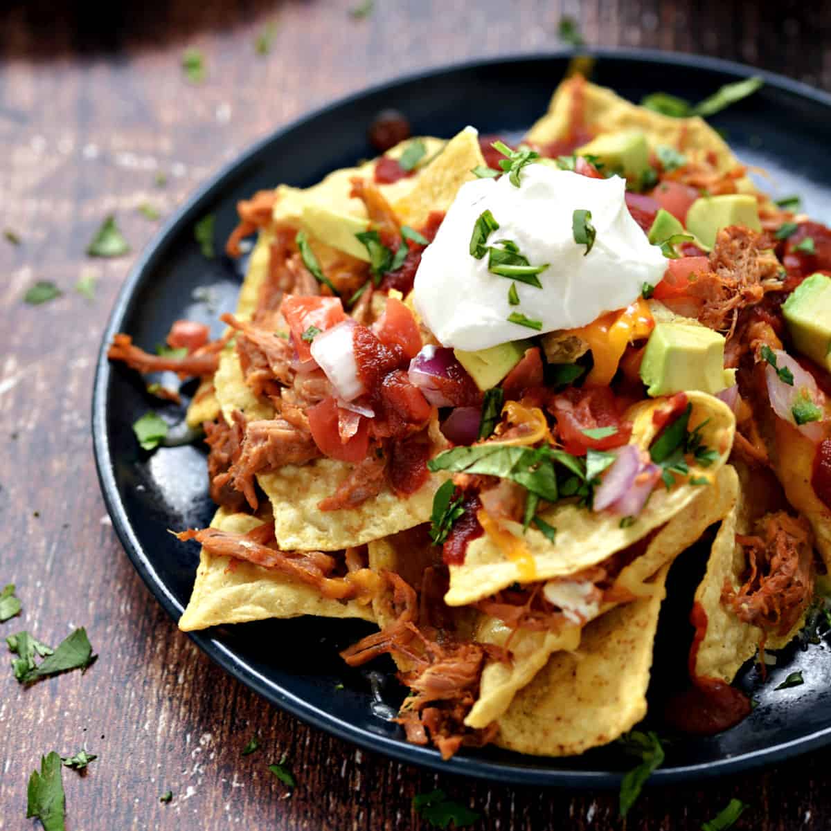Tortilla chips, melted cheese, pulled pork, pico de gallo, avocado chunks, sour cream and cilantro piled up on a blue plate.