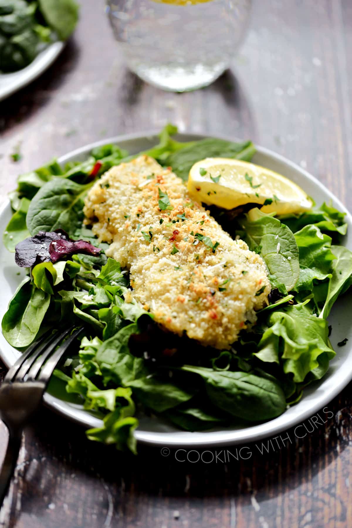 Oven Baked Cod with Panko Crumbs served on a bed of leafy greens with a lemon wedge on the side and sparking water with lemon in the background.