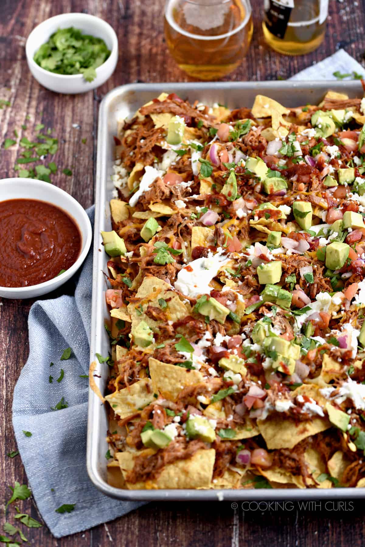 Tortilla chips, pulled pork, melted cheese, diced avocado, sour cream and barbecue sauce in a baking sheet with bowls of barbecue sauce, and chopped cilantro in the background.