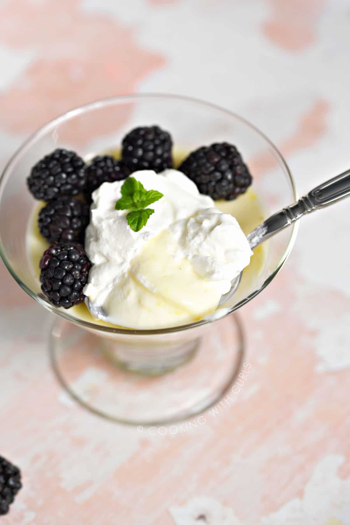 A spoon pulling up a scoop of lemon posset topped with whipped cream, with fresh blackberries around the left side of the dessert glass.