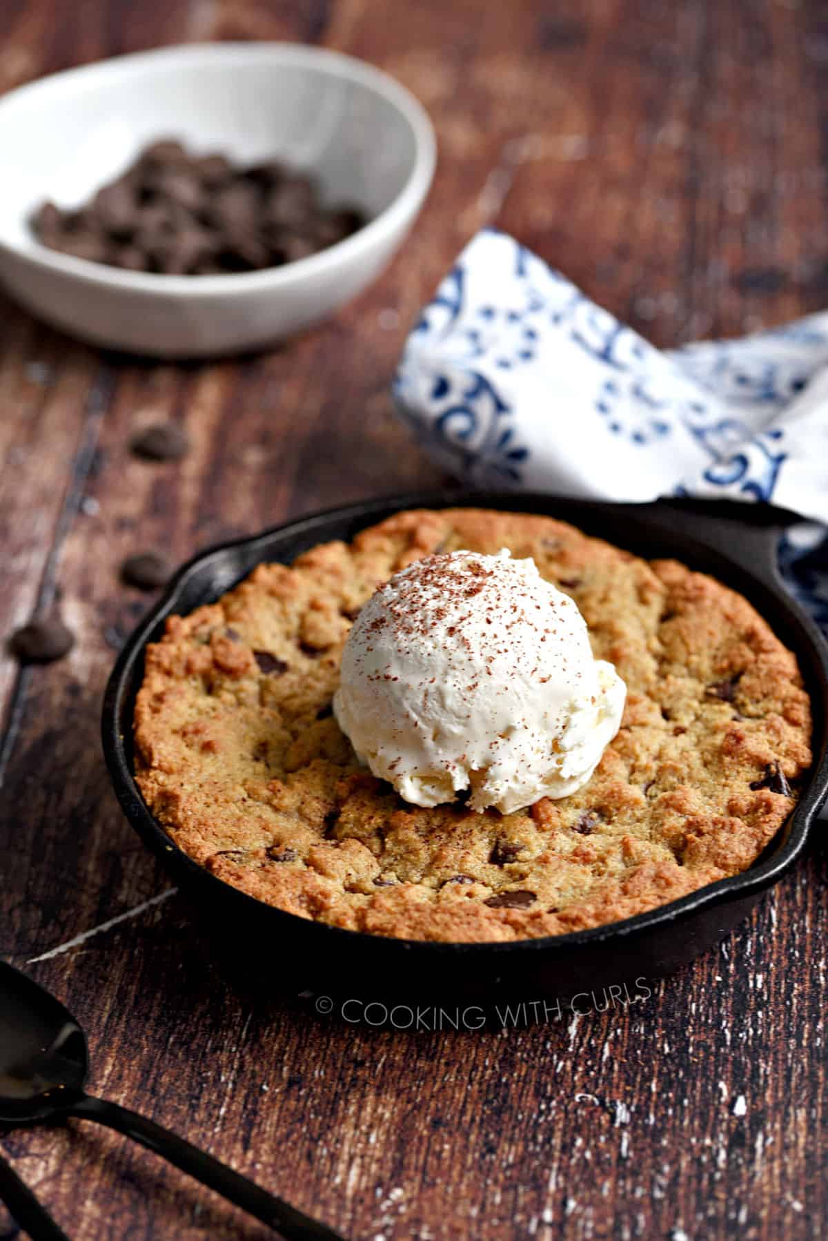 Mini cast iron skillet with a chocolate chip cookie topped with a scoop of vanilla ice cream, with a white bowl of chocolate chips in the background.
