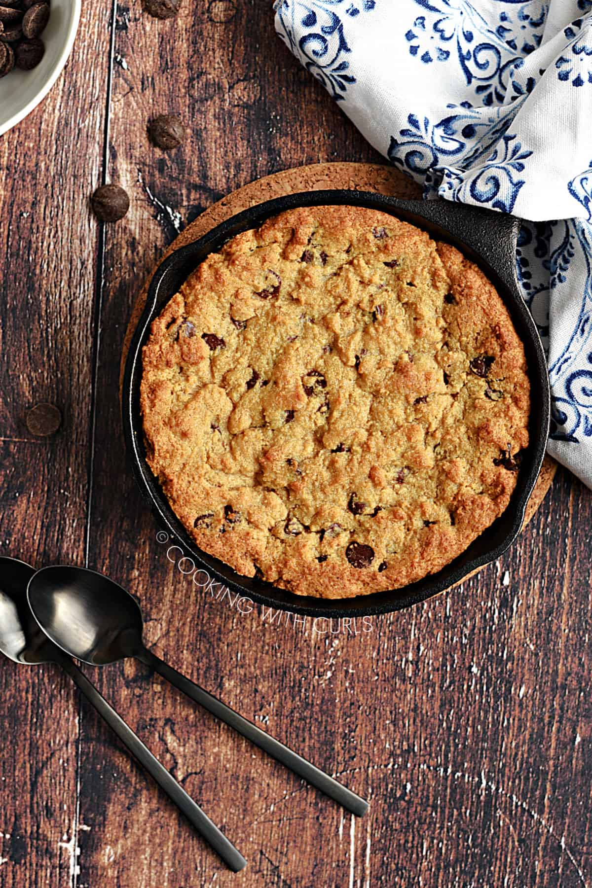 Looking down over a mini skillet chocolate chip cookie with two black spoons and scattered chocolate chips.
