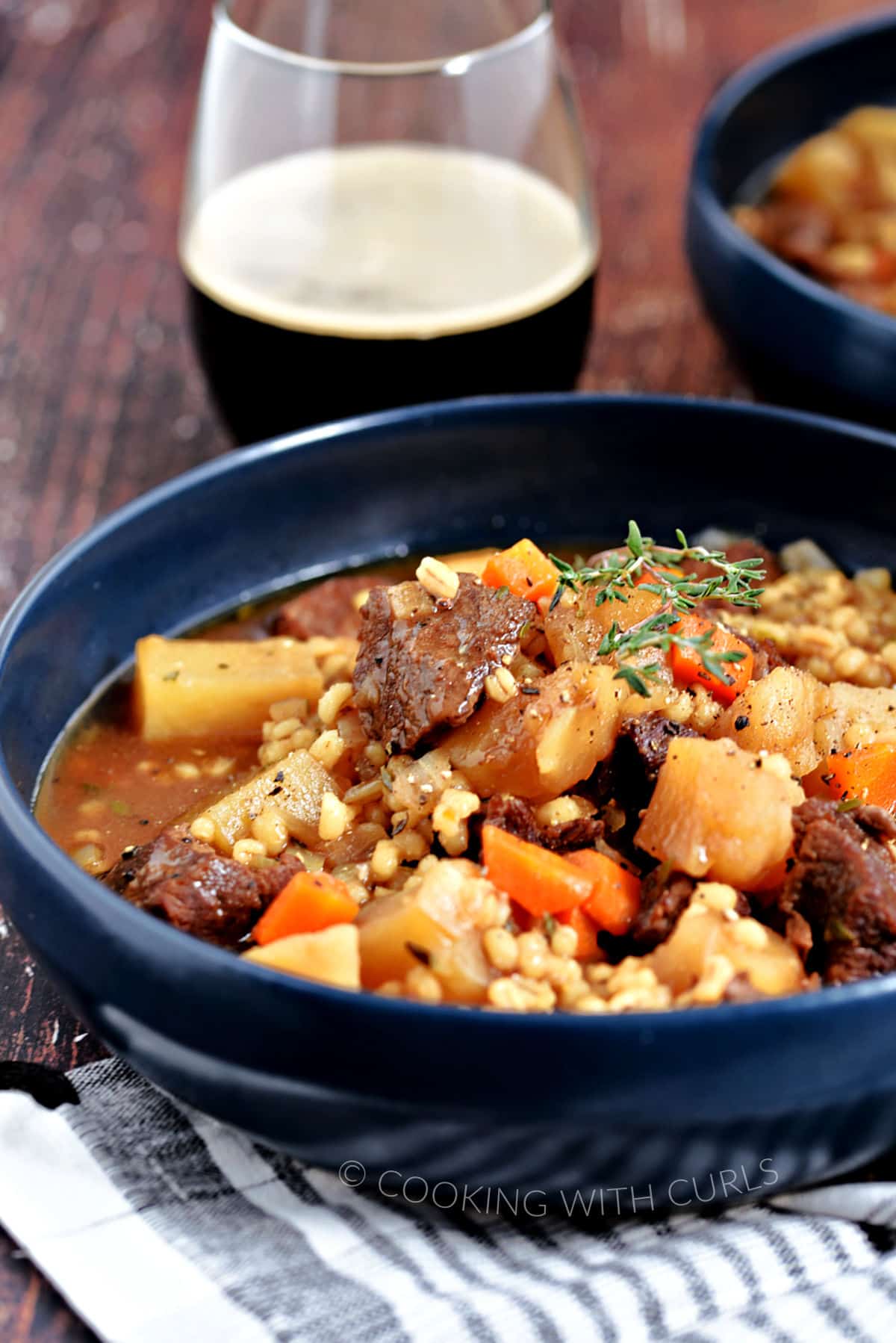 Large chunks of beef, potatoes and carrots surrounded by barley and broth in a blue bowl with a glass of Guinness in the background.