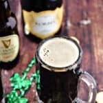Foamy, black cocktail in a glass beer mug surrounded by green beads with bottles of Guinness and champagne in the background.